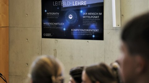 Im Vordergrund unscharfe Silhouetten der Zuhörer, im Hintergrund im Fokus eine Tafel mit dem Titel: Leitbild Lehre