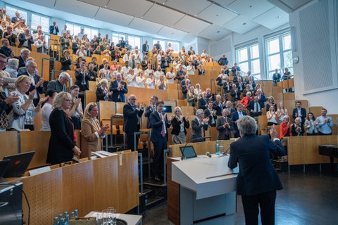 Hörsaa-Perspektive von unten. Vorne unten steht der Vortragende, die Zuhörende stehen vor ihren Plätzen und applaudieren. 
