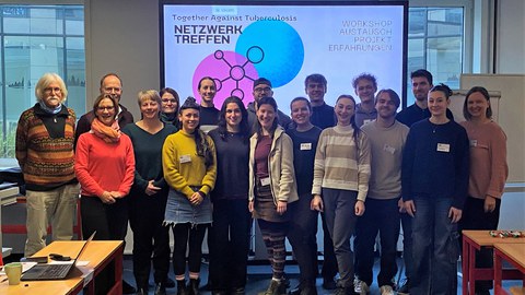 Eine Gruppe Menschen steht in einem Seminarraum vor einem großen Bildschirm mit der Überschrift "Together against Tuberculosis - Netzwerktreffen"