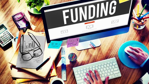 Ein Schreibtisch, auf dem ein Computer, eine Tasse Kaffee, ein Taschenrechner, eine Brille, Hefte und Dokumente sowie Stifte liegen.