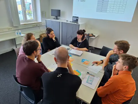 Sieben Menschen sitzen am Tisch, vor ihnen Blätter mit EKG und Zettel mit verschiedenen Farben.