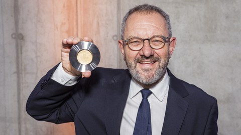 Ein lächelnder Mann zeigt in die Kamera eine Medaille in seiner Hand.