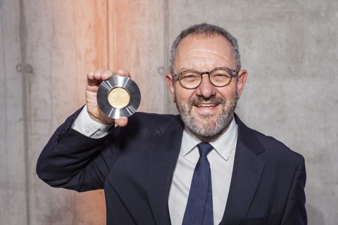 Ein lächelnder Mann zeigt in die Kamera eine Medaille in seiner Hand.