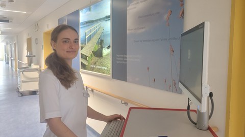 Eine junge Frau in einem weißen Kittel steht vor einem PC-Bildschirm in einem Krankenhausflur.