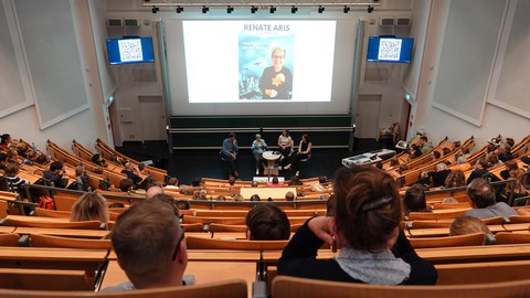 Menschen sitzen in einem vollen Hörsaal. Auf der Leinwand ein Foto von Renate Aris mit ihrem Namen als Überschrift.