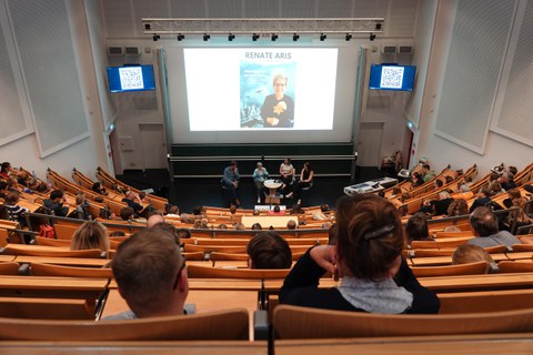 Menschen sitzen in einem vollen Hörsaal. Auf der Leinwand ein Foto von Renate Aris mit ihrem Namen als Überschrift.