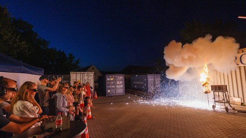 Links eine Gruppe von Zuschauern, die Schutzbrillen und Gehörschutz tragen, rechts eine brennende Puppe, die helle Funken herum fallen lässt.