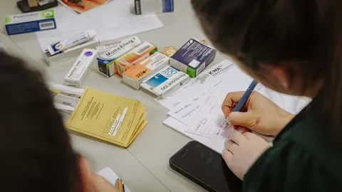 Über die Schulter von zwei Personen fokussiert die Kamera auf den Tisch, auf dem Impfausweise, Medikamente und Formulare für hausärztliche Verschreibungen liegen.