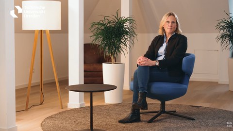 Links stehen Zimmerelemente: Eine Stehlampe, ein Kaffeetisch, ein Pflanzentopf. Rechts: Eine Person sitzt in einem Stuhl 