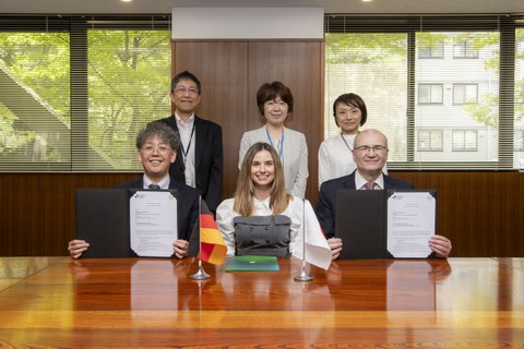 Drei Personen sitzen am Tisch, drei weitere Personen stehen hinter ihnen. Auf dem Tisch stehen die Flaggen von Japan und Deutschland. Die Personen links und rechts am Tisch halten jeweils ein unterzeichnetes Dokument in die Kamera.