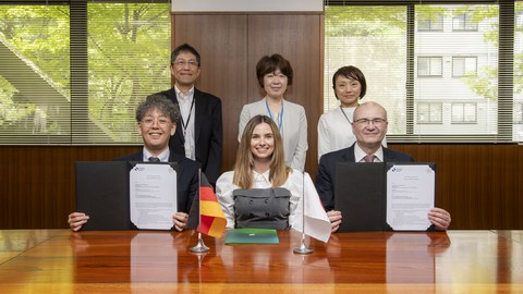 Drei Personen sitzen am Tisch, drei weitere Personen stehen hinter ihnen. Auf dem Tisch stehen die Flaggen von Japan und Deutschland. Die Personen links und rechts am Tisch halten jeweils ein unterzeichnetes Dokument in die Kamera.