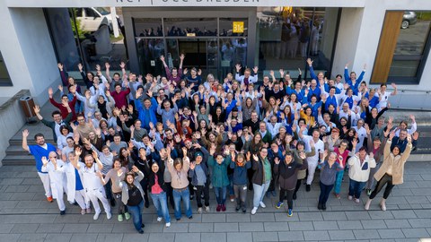 Eine große Gruppe von Menschen winkt in die Kamera vor einem Gebäude. Am Gebäude Schriftzug "NCT | UCC Dresden".