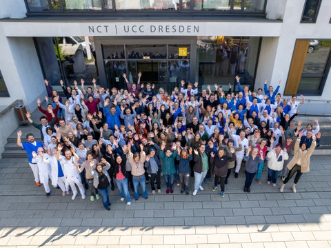 Eine große Gruppe von Menschen winkt in die Kamera vor einem Gebäude. Am Gebäude Schriftzug "NCT | UCC Dresden".