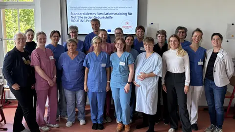Eine Gruppe von Menschen, einige davon in Kassacks, steht vor einem Bildschirm und schaut in  die Kamera.