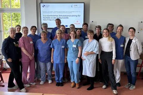Eine Gruppe von Menschen, einige davon in Kassacks, steht vor einem Bildschirm und schaut in  die Kamera.