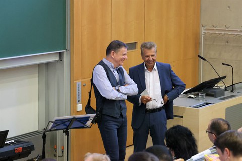 Zwei Männer stehen in einem Hörsaal vor dem beginn einer Veranstaltung und lachen.