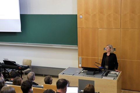Eine Frau steht an einem Hörsaal-Pult und spricht vor vielen Zuhärenden. Links von ihr ist eine leere große Tafel.