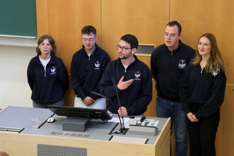 Fünf junge Menschen in glaichfarbigen Kapuzen-Pullovern stehen am Pult eines Hörsaals. Sie sprechen vor einem Auditorium.  