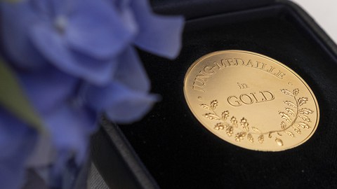 Eine Jung-Medaille in Gold auf schwarzem Hintergrund, im Vordergrund links unfokussiert lila Blumen.