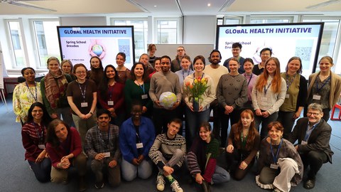 Rund 30 Menschen posieren vor der Kamera. Im Hintergrund zwei Bildschirme mit dem Text "Global Health Initiative"