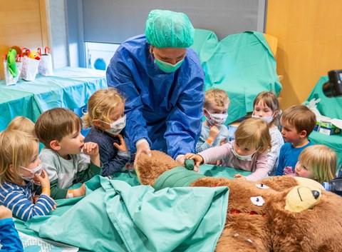 Ein Teddybär liegt auf einem OP-Tisch. Eine Person in OP-kleidung hält das Bein des Bären. Rund um den Tisch stehen kleine Kinder, eins davon bindet einen Verband um das Bein.
