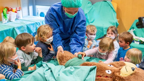 Ein Teddybär liegt auf einem OP-Tisch. Eine Person in OP-kleidung hält das Bein des Bären. Rund um den Tisch stehen kleine Kinder, eins davon bindet einen Verband um das Bein.