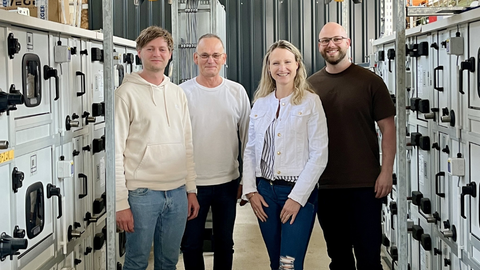 Vier Menschen stehen in einer industriellen Anlage in einem Gang zwischen zwei Reihen von Minicontainern mit Türen.