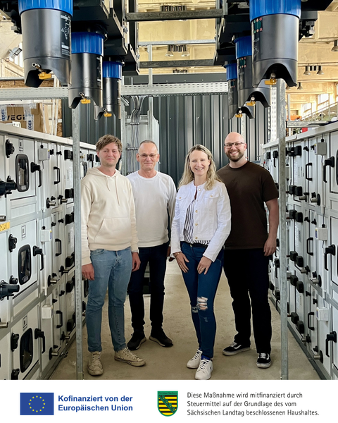 Vier Menschen stehen in einer industriellen Anlage in einem Gang zwischen zwei Reihen von Minicontainern mit Türen.