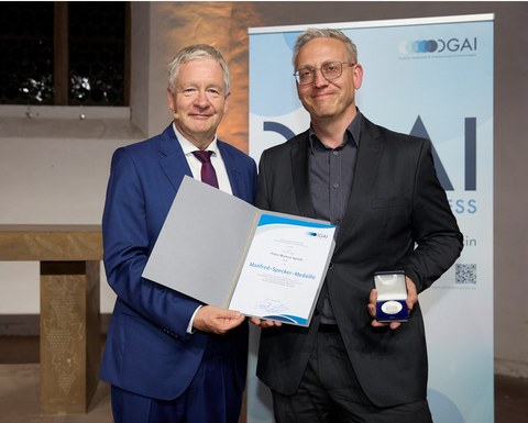 Zwei festlich gekleidete Personen stehen nebeneinander, halten eine Urkunde und eine Medaille und lächeln in die Kamera. 