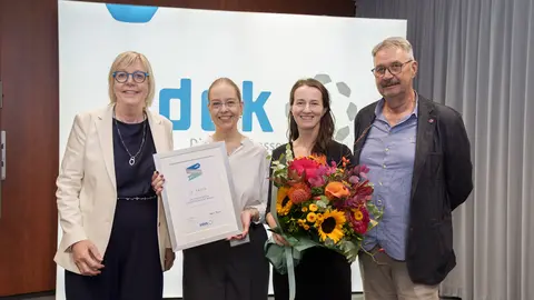 Vier Personen stehen vor einer weißen Tafel mit teilweise sichtbarer Überschrift "vdek", eine Person hält eine Urkunde, eine andere Person hält einen Blumenstrauß.
