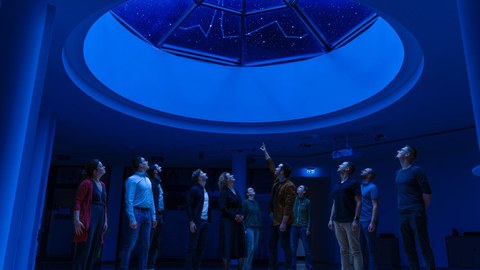 Eine Gruppe von Menschen steht in einem dunklen Raum unter einem runden Dachfenster; durch das Fensterglas ist ein Sternbild sichtbar, das an die Pulslinie erinnert.