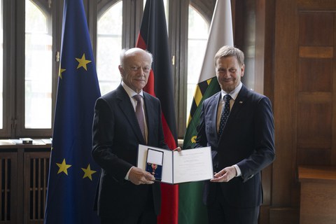 Auf dem Bild sind zwei Männer zu sehen, die gemeinsam eine Verleihungsurkunde mit einem Orden halten. Im Hintergrund stehen die Fahnen von Sachsen, Deutschland und der EU.