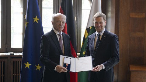 Auf dem Bild sind zwei Männer zu sehen, die gemeinsam eine Verleihungsurkunde mit einem Orden halten. Im Hintergrund stehen die Fahnen von Sachsen, Deutschland und der EU.