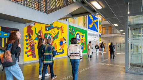 Foto einer Gruppe Studierender von hinten, die durch einen buntbemalten Gang auf dem TUD-Campus laufen.