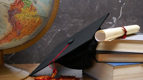 Nahaufnahme von einem Tisch. Links ist ein Teil vom Globus mit Südamerika sichtbar, rechts gestapelt drei Bücher und darauf eine gerollte Urkunde, links an der Buchstapel hängt ein Akademikerhut.