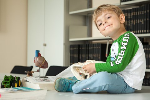 A young boy sits among open medical books, a heart model and a toy tractor on the table in the library, playing with a model of a human skull.
