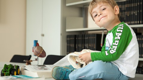 Ein kleiner Junge sitzt zwischen aufgeschlagenen Medizinbüchern, einem Herzmodell und einem Spielzeugtraktor auf dem Tisch in der Bibliothek und spielt mit dem Modell eines menschlichen Schädels.