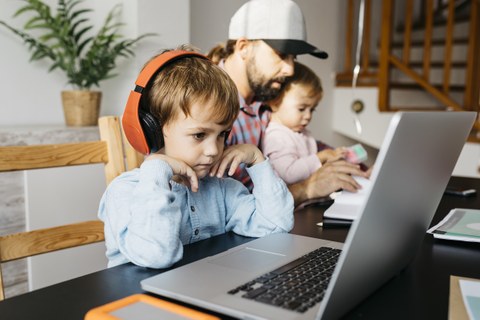 Foto eines Vaters mit seinen zwei kleinen Kindern im Homeoffice. Der ältere sitzt mit Kopfhörern am Laptop.