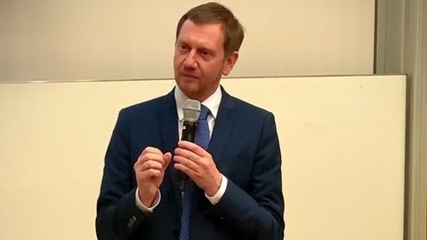 Sachsens Ministerpräsident Kretschmer steht mit einem Mikrofon in der Hand vor einer weißen Wand. Er blickt an der Kamera vorbei.