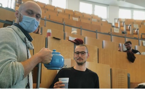 3 students in an auditorium smile at the camera. One is being served tea.