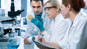 Das Foto zeigt drei Personen in weißen Laborkitteln. Sie sitzen vor einem Mikroskop. Eine der Personen hält eine Petrischale mit Bakterien in der Hand. Eine andere Person macht Notizen auf einem Block.