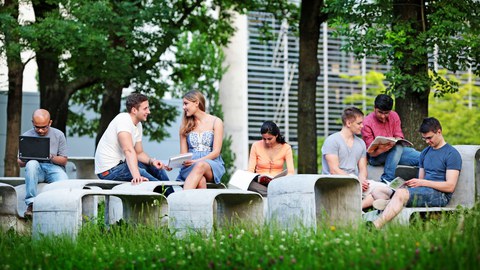 Das Foto zeigt mehrere Studierende draußen im Grünen. Sie sitzen auf einer geschwungenen Betonformation aus Tischen und Bänken und lernen.