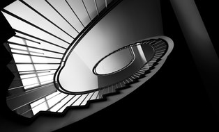 Treppe im Andreas-Schubert-Bau. Das Foto wurde vom Boden aus aufgenommen und ist in schwarz-weiß. Aus dieser Perspektive verläuft die Treppe spiralförmig nach oben.