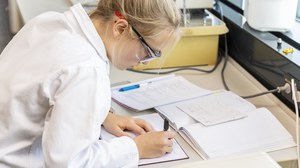 Foto einer Frau, die im Labor an einem Schreibtisch sitzt und sich etwas in ein Heft notiert