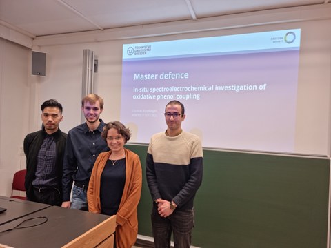 Drei Männer und eine Frau stehen vor einer Präsentation mit dem Titel 'Master Defence In Situ Spectroelectrochemical investigations of oxidative phenol coupling'