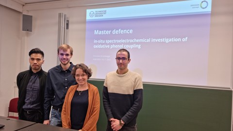 Drei Männer und eine Frau stehen vor einer Präsentation mit dem Titel 'Master Defence In Situ Spectroelectrochemical investigations of oxidative phenol coupling'