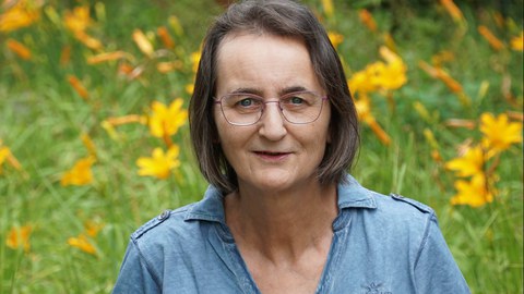 Frau mit Brille und braun-grauen Haaren in blauem Oberteil vor einem Blumen-Hintergrund