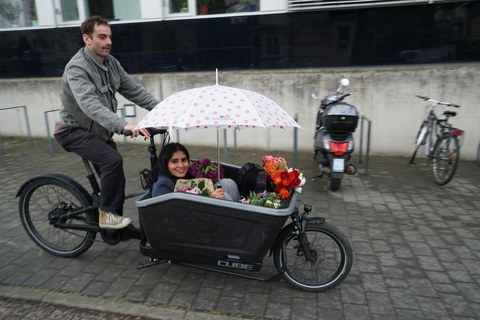 Ein Mann fährt ein Lastenrad in dem eine Frau sitzt die einen Regenschirm hält. In dem Lastenrad sind auch einige Blumensträuße.