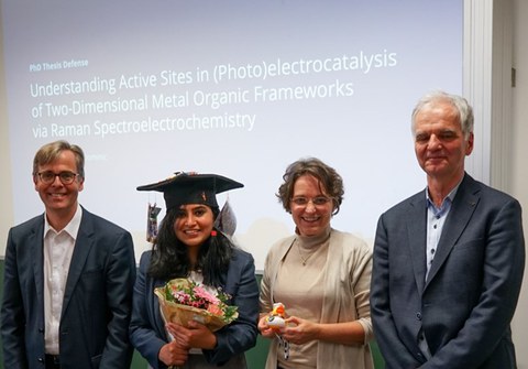 Vier Personen (von links nach rechts: Mann, Frau Mit Doktorhut und Blumen, Frau, Mann) stehen vor einer Präsentation auf der steht "PhD Thesis Defense understanding active sites in (photo)electrocatalysis of two-dimensional metal organic frameworks via Raman spectroelectrochemistry