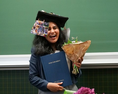 Eine Frau mit doktorhut lacht und hält ein Buch mit der Aufschrigt Doktorarbeit sowie einen Blumenstrauß